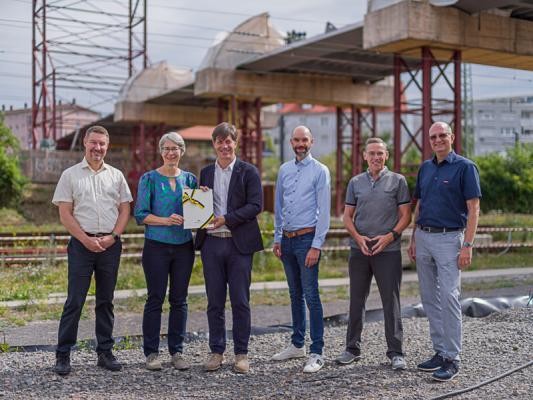 Staatssekretärin Elke Zimmer (2. v. l.) hat den Förderbescheid des Landes Baden-Württemberg in Höhe von 13,75 Millionen Euro am Freitag, 26. Juli 2024, an Jürgen Odszuck, Erster Bürgermeister der Stadt Heidelberg überreicht. Mit im Bild: Klaus-Peter Hofbauer, Leiter des Tiefbauamts, Gerald Dietz, Leiter Geschäftsstelle Bahnstadt, Christian Hemkendreis, DSK GmbH (v. r.) sowie Kai Zumkeller, leitender Baudirektor im Regierungspräsidium Karlsruhe (l.). (Foto: Dittmer) Sechs Personen posieren mit einer Urkunde vor dem Rohbau einer Brücke