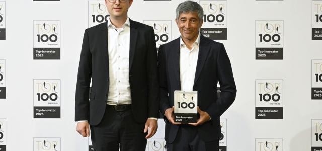 Zwei Männer stehen vor der Kamera. Einer von ihnen hält eine Skulptur, auf der eine 100 zu sehen ist.