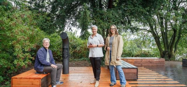 Ein Mann und zwei Frauen stehen auf einer neu errichteten Fläche aus Holz mit Sitzbänken und einem periskopartigen Gerät