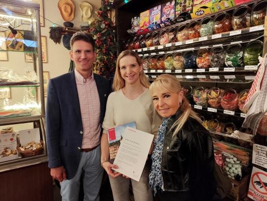 Marc Massoth (l.) und Iris Schiller (r.) von der Wirtschaftsförderung übergaben ein Schreiben des Oberbürgermeisters an Antonia Brehme, Inhaberin des Zuckerladens. (Foto: Stadt HD) Ein Mann und zwei Frauen lächeln in die Kamera, die Frau in der Mitte hält ein Schriftstück in den Händen.
