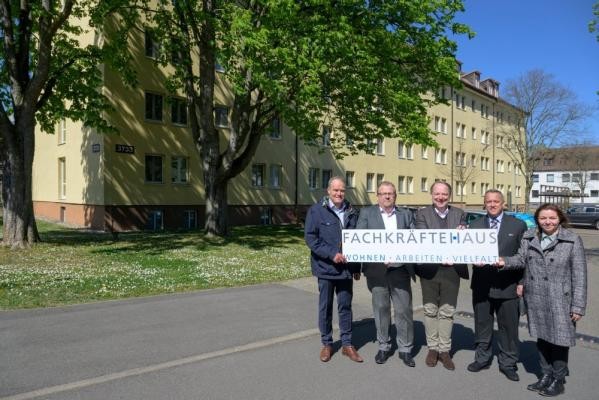 Vor dem neuen Fachkräftehaus (v.l.): Oberbürgermeister Eckart Würzner, Gerald Kraus, Prokurist bei der Gesellschaft für Grund- und Hausbesitz mbH Heidelberg (GGH), Steffen Grimm, Leiter des Bereichs Personal bei der Rhein-Neckar-Verkehr GmbH (rnv), Christos Roufakis, einer der ersten Bewohner und Busfahrer bei der rnv, und Nadine Hülden, Geschäftsführerin der Heidelberger Dienste. (Foto: Rothe) Gruppenfoto mit vier Männern und einer Frau die einen Banner mit der Aufschrift "Fachkräftehaus" vor sich halten. Im Hintergrund ist ein mehrstöckiges Haus zu sehen.
