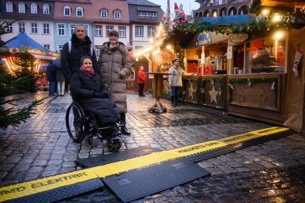 Hilfe für den barrierefreien Weihnachtsmarktbesuch: Das Ehepaar Förster und Nadine Layer(r.) vom Team der Kommunalen Behindertenbeauftragten testeten die neuen Kabelbrücken-Rampen beim Weihnachtsmarkt vor dem Heidelberger Rathaus. (Foto. Nicolaus Niebylski) Zu sehen sind 3 Personen auf dem Weihnachtsmarkt. Ein Frau sitzt im Rollstuhl und steht vor einer Rampe über eine Kabelbrücke.