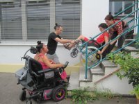 Zwei Personen transportieren eine Person im Rollstuhl eine Treppe heraus, eine weitere beobachtet