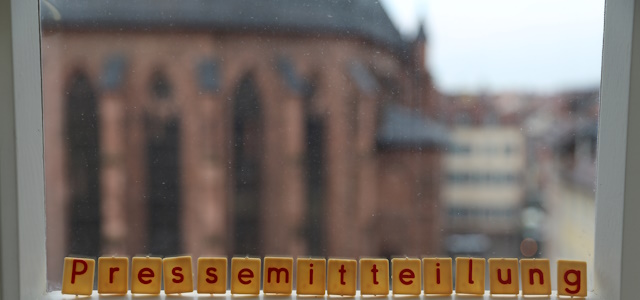 Das Wort "Pressemitteilung" in Scrabble Buchstaben auf eine Fensterbank gelegt. Die Heiliggeistkirche ist im Hintergrund zu sehen.