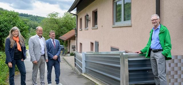 Gruppenfoto vor Haus. Elemente der Schutzwand aus Metall liegen gestapelt an der Hauswand