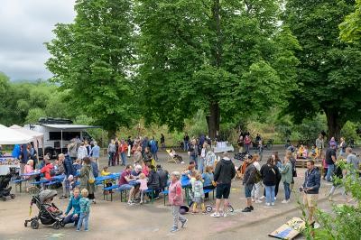 Menschen sitzen auf Biertischbänken auf einem Platz mit großen Bäumen