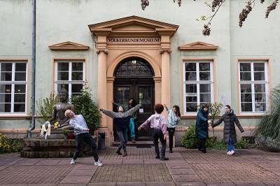 Sechs Personen in Freizeitkleidung tanzen vor dem Eingang des Museums